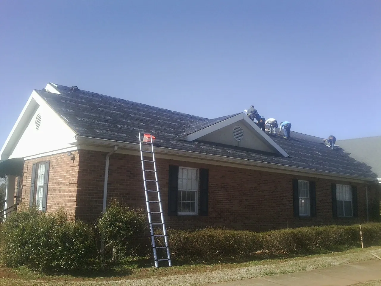 Crew tearing shingles off a brick ranch roof with a ladder set — Emergency Roof Repair work by our crew in Keene