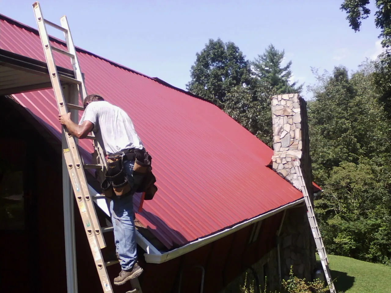 Roofer on a ladder beside a red metal roof and stone chimney — Soffit & Fascia Repair by Roofing Brothers in Keene